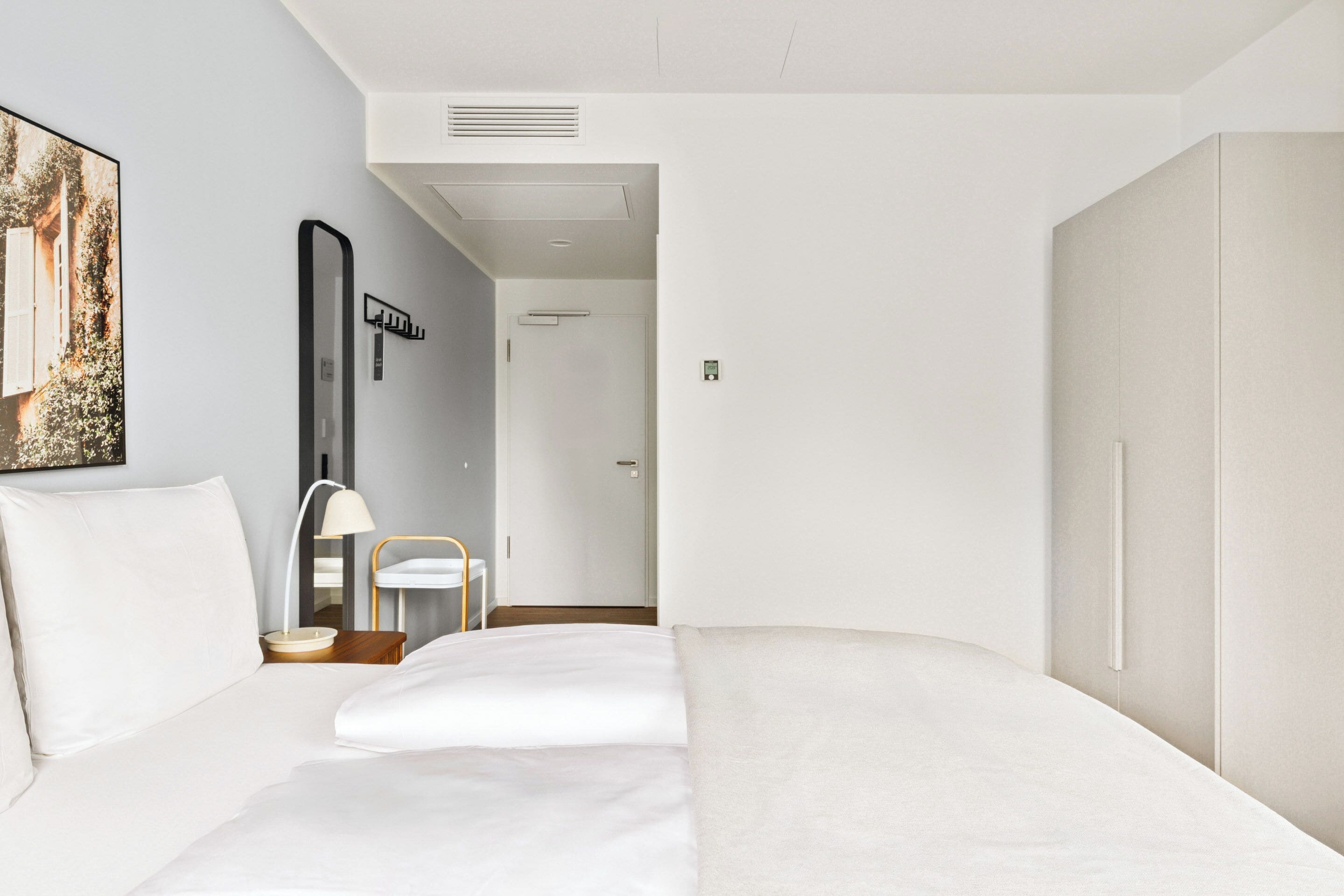A modern hotel room with a large bed, a bedside table with a lamp, a chair, and a door leading to another room.