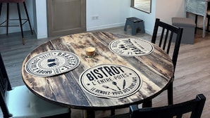 A dining area with a wooden table, four chairs, and a round tablecloth with a logo. There is a door with a phone and a small stool near it. - Cosy and relaxing atmosphere in St Zacharie, in the heart of the village. (Unknown)