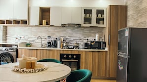 A modern kitchen with wooden cabinets, a built-in oven, a refrigerator, and a dining area with a round table and teal chairs. - The Snug Haven (Nairobi)