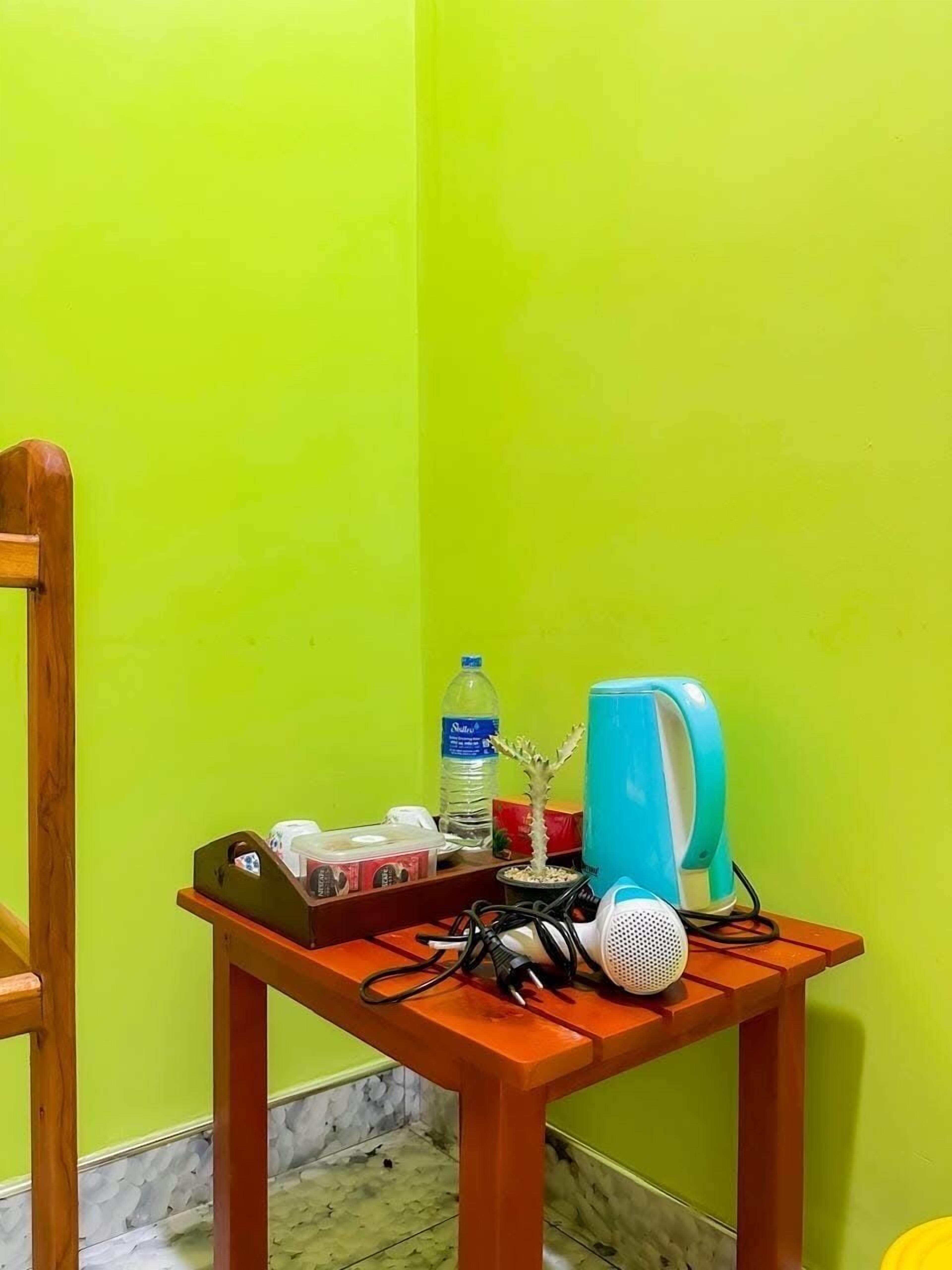 Une petite table en bois, sur laquelle on aperçoit un haut-parleur bleu, une bouteille d’eau et quelques petits objets, le tout placé devant un mur vert vif.