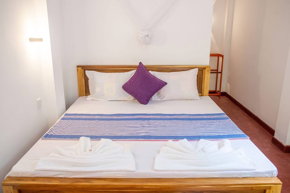A neatly made bed with a wooden headboard, two white pillows, a purple accent pillow, and a striped blanket. There are two neatly folded towels on the bed.