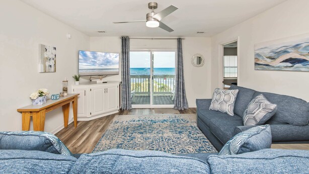 A living room with a sofa, a TV, a ceiling fan, a view through a glass door to the outside, and a painting on the wall. - Serenity is Yours! Oceanfront Pool,SP3 (North Myrtle Beach)
