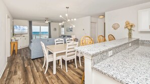 A modern kitchen with granite countertops, a dining area with white chairs, and a view of the ocean. - Serenity is Yours! Oceanfront Pool,SP3 (North Myrtle Beach)
