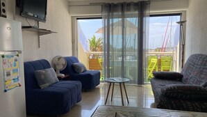 A living room with a blue sofa, a patterned armchair, a small table, and a large sliding glass door leading to a balcony with a view of boats and a building. - Charming studio in enjoyable La Grande-Motte with WiFi (La Grande-Motte)