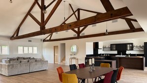 A spacious living area with a high ceiling and exposed wooden beams, a dining table with colorful chairs, and a modern kitchen in the background.