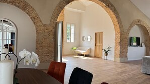 A spacious room with high ceilings and exposed brick arches, a dining area with a wooden table and chairs, and a cozy seating area with a sofa and a TV.