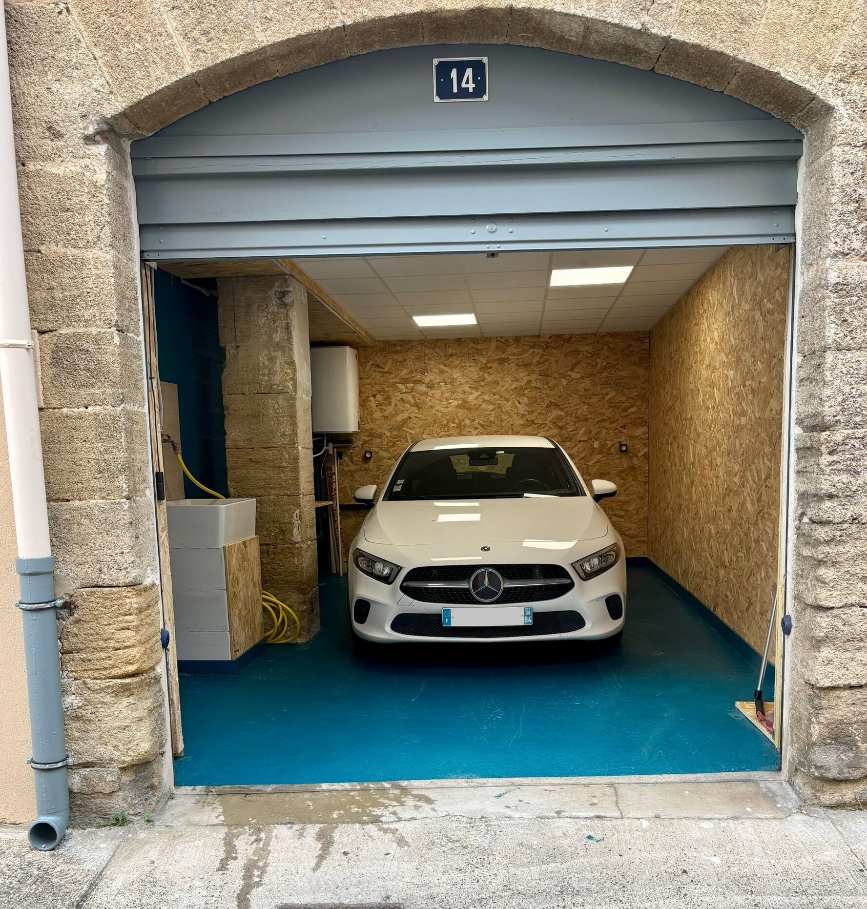 A white car parked inside a garage with a blue floor, a stone wall, and a sign indicating the number 14.