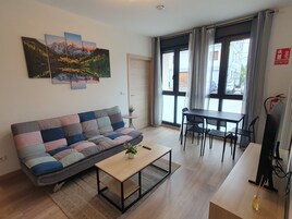 Una sala de estar con un sofá, una mesa de centro, una mesa de comedor con sillas, una planta en maceta y un cuadro en la pared.
