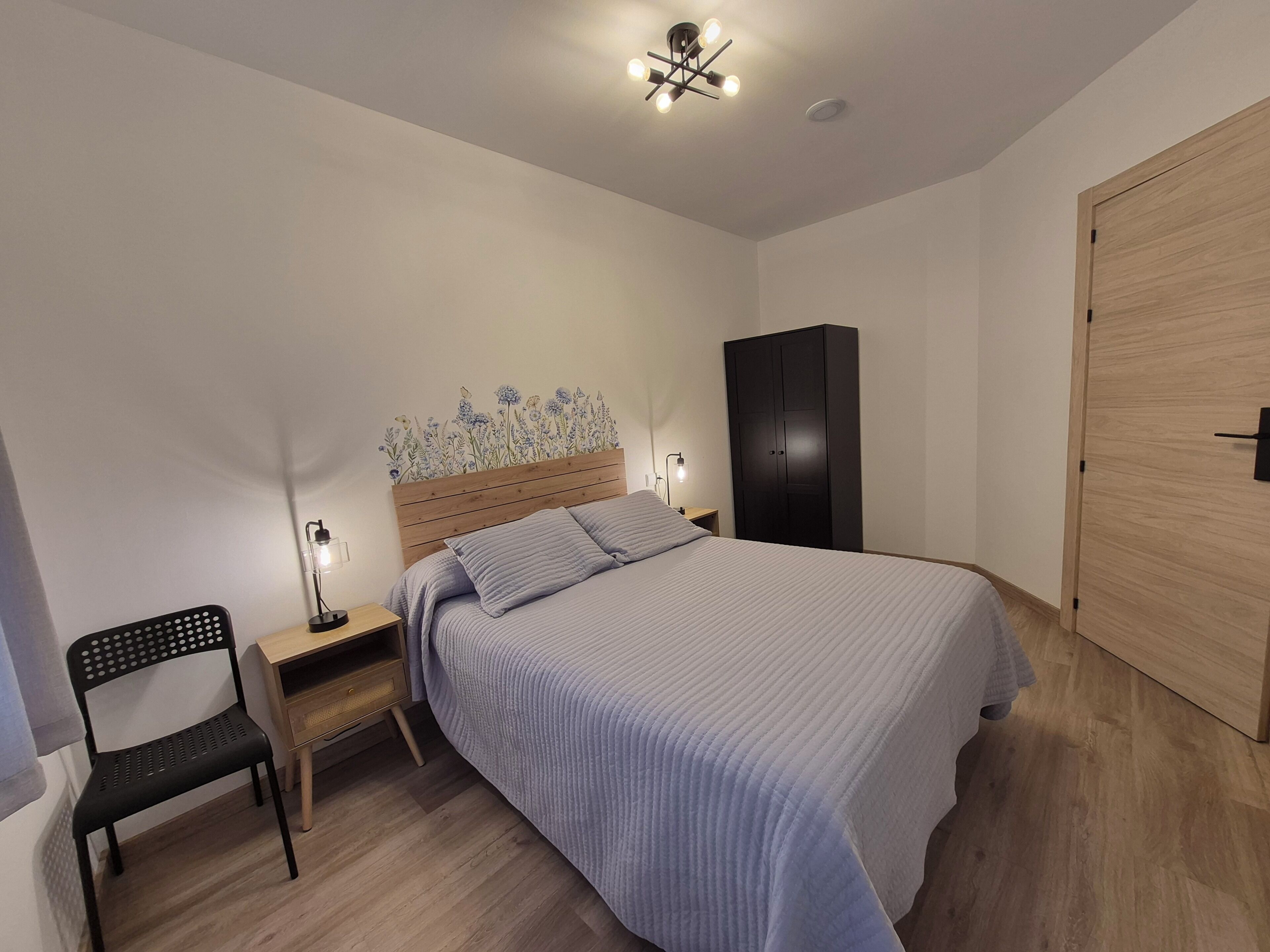 Un dormitorio con una cama, una mesita de noche, una silla, un armario y una pared con papel tapiz floral.