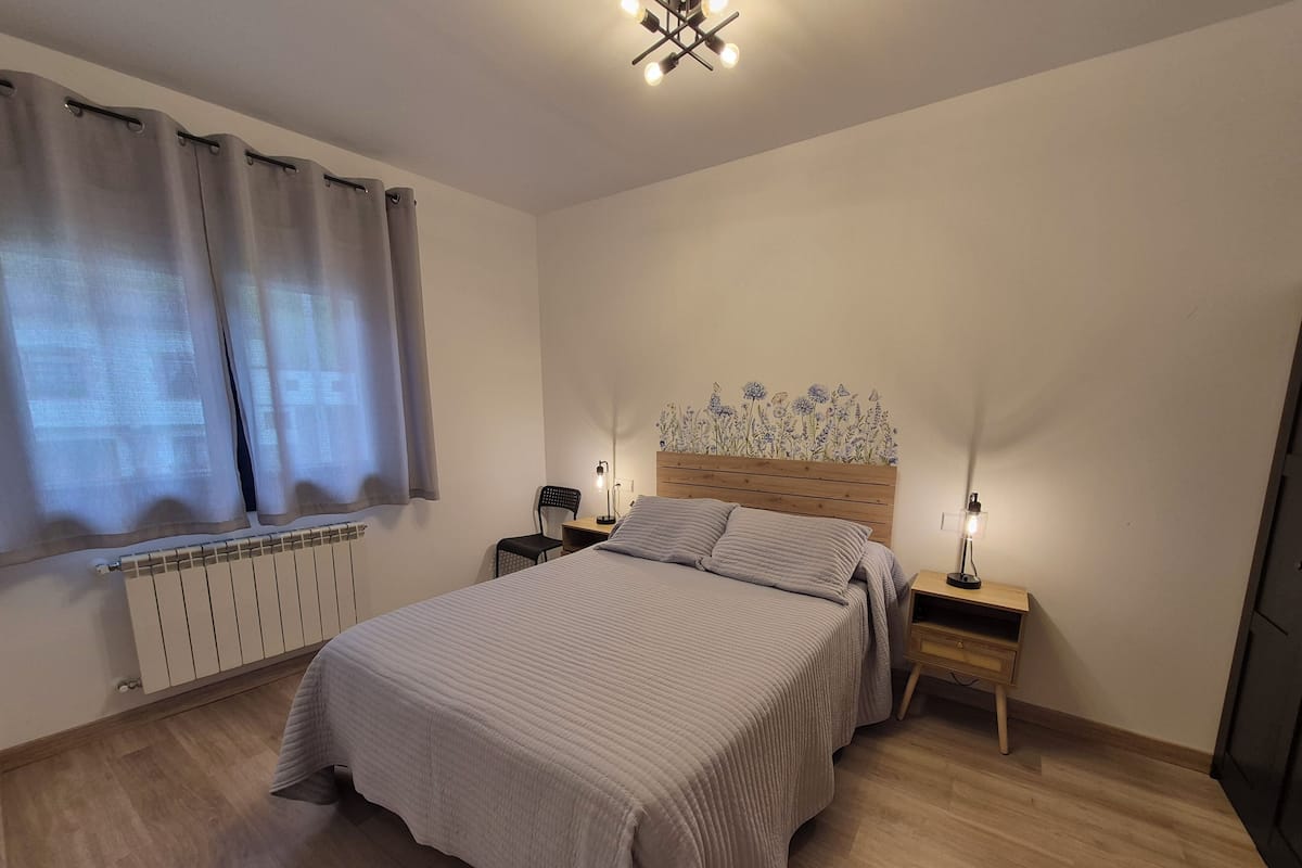 Een slaapkamer met een bed, nachtkastjes, een stoel, een radiator en een raam met gordijnen.