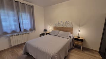 Un dormitorio con cama, mesitas de noche, una silla, un radiador y una ventana con cortinas.