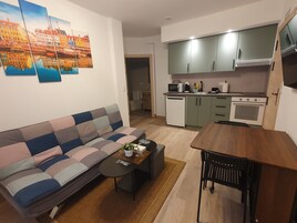 Un espacio habitable compacto con sofá, una pequeña mesa de comedor, zona de cocina con microondas, lavavajillas y armarios.