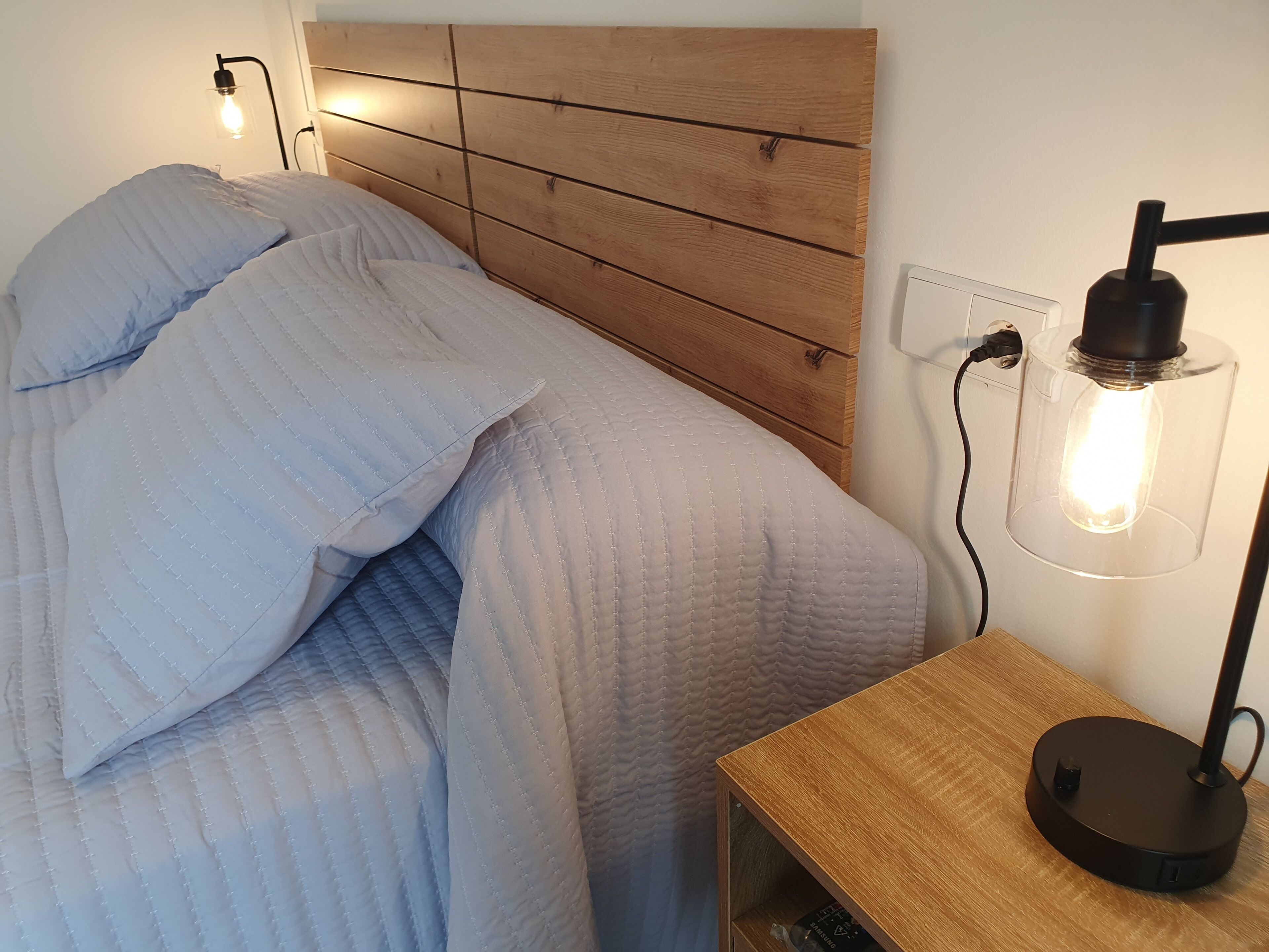 Una cama con cabecera de madera, una mesita de noche con lámpara y un enchufe de pared.