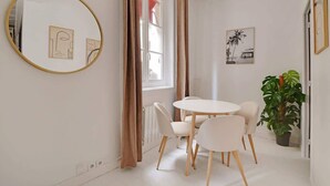 A modern, minimalist room with a round mirror, a white table with four chairs, a potted plant, and two framed artworks on the walls.
