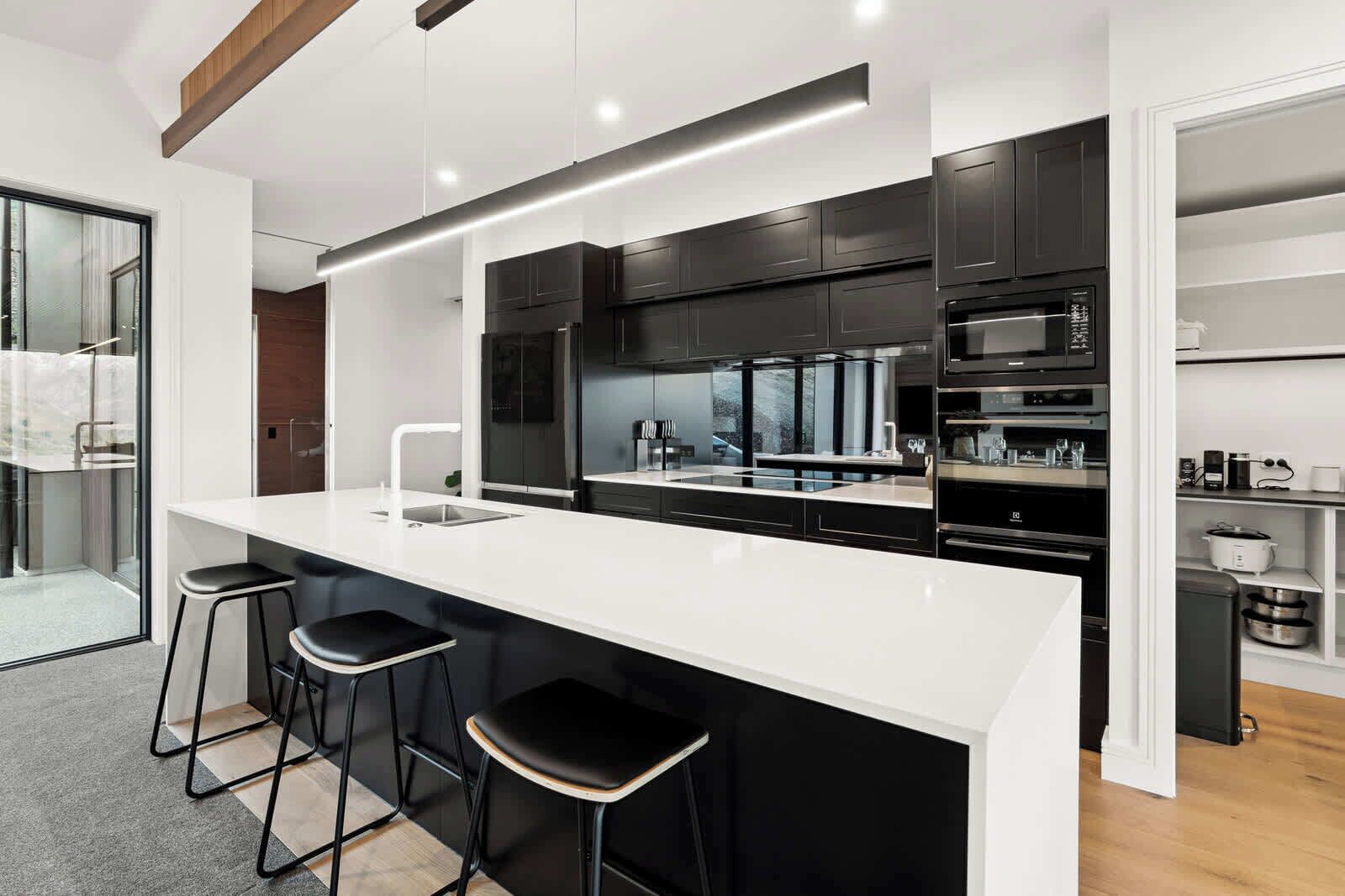 Una cocina moderna con encimera blanca, armarios negros y electrodomésticos de acero inoxidable. Hay tres taburetes de bar y un ventanal que proporciona luz natural.