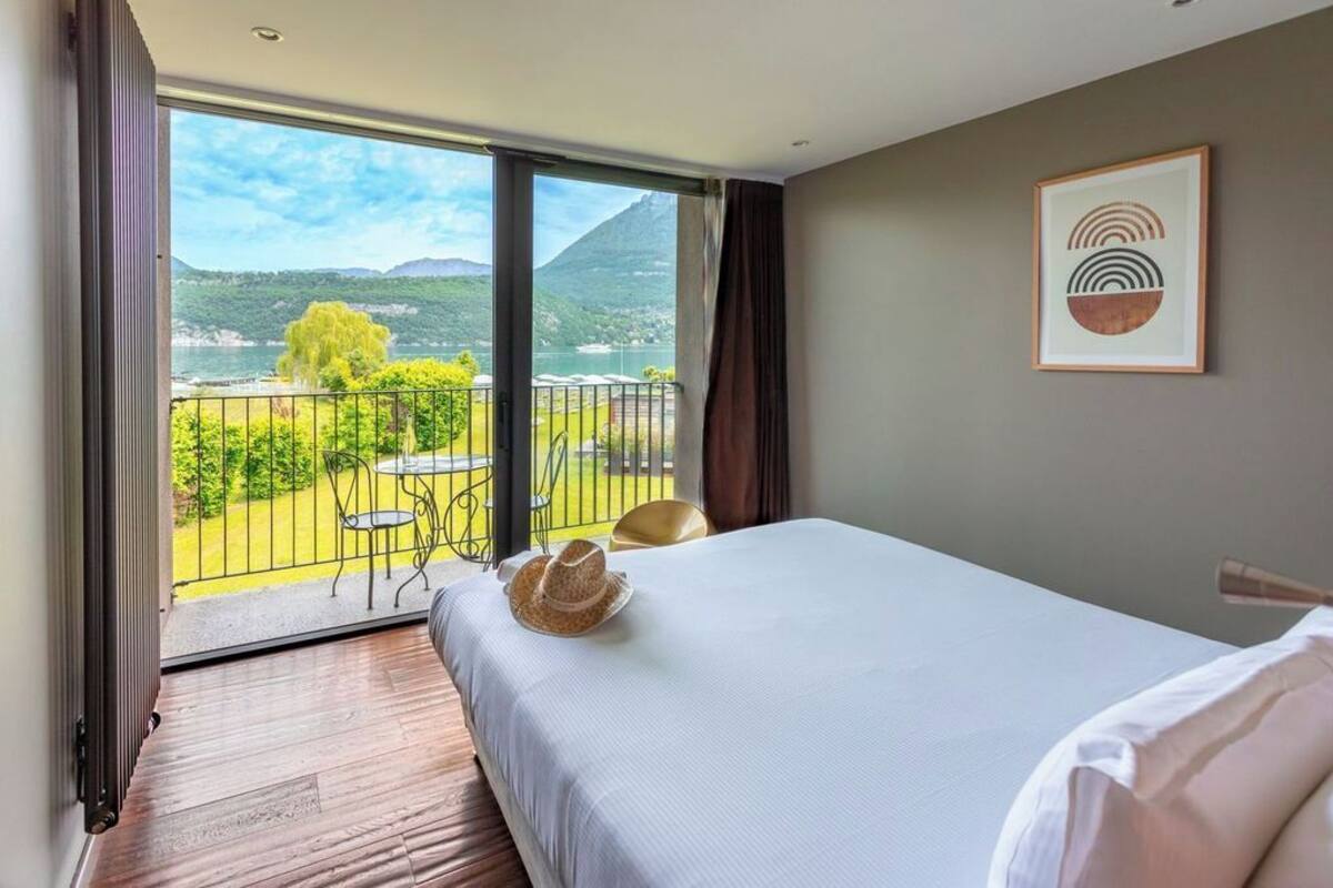 A hotel room with a bed, a view of a balcony with outdoor furniture, and a framed artwork on the wall.