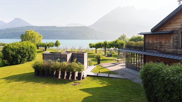 A lakeside property with a wooden cabin, a patio with seating, and a well-maintained lawn.