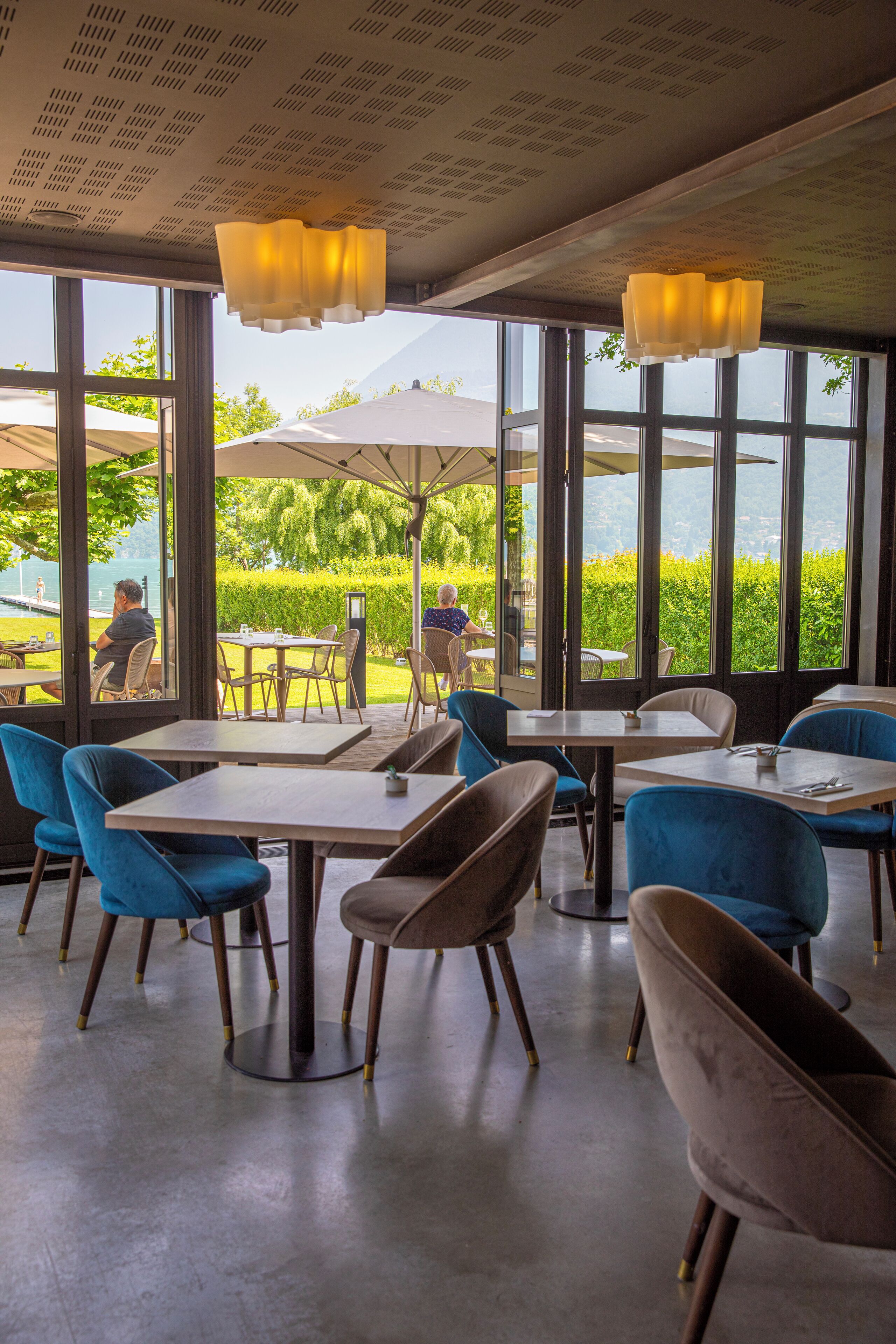 An indoor dining area with tables and chairs, large windows with a view of greenery and a mountain, and hanging pendant lights.