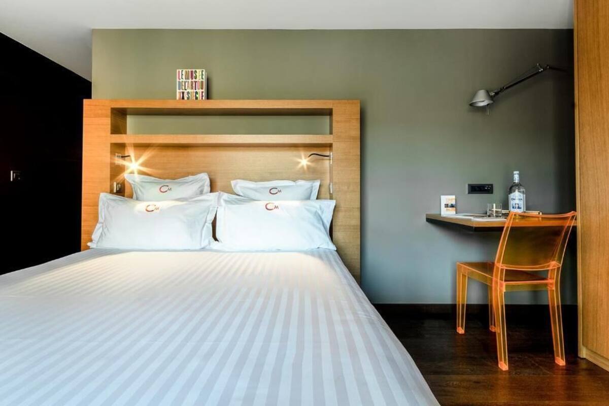 A hotel room with a large bed, a desk with a chair, a lamp, and a bottle on the desk.