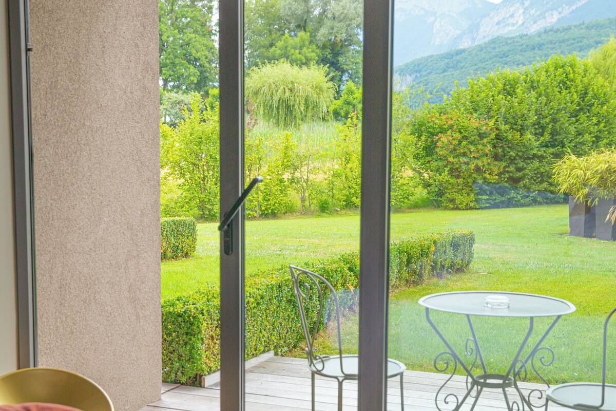 A bedroom with a large sliding glass door leading to a balcony with a table and chairs, a bed with a rolled-up towel, and a view of a garden and mountains.