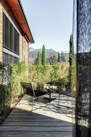 A wooden deck with outdoor furniture, a view of mountains, and a building with a sloped roof and shutters.