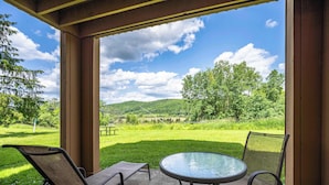 Un porche techado con una mesa de vidrio y dos sillones, con vistas a una zona verde con árboles y colinas.
