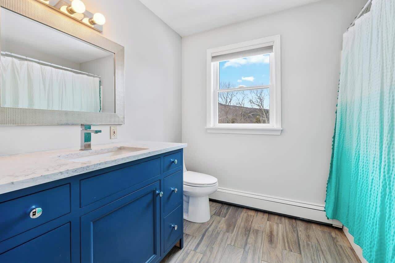 Un bagno con un WC bianco, un mobile lavabo blu, uno specchio grande con illuminazione e una finestra vista alberi.