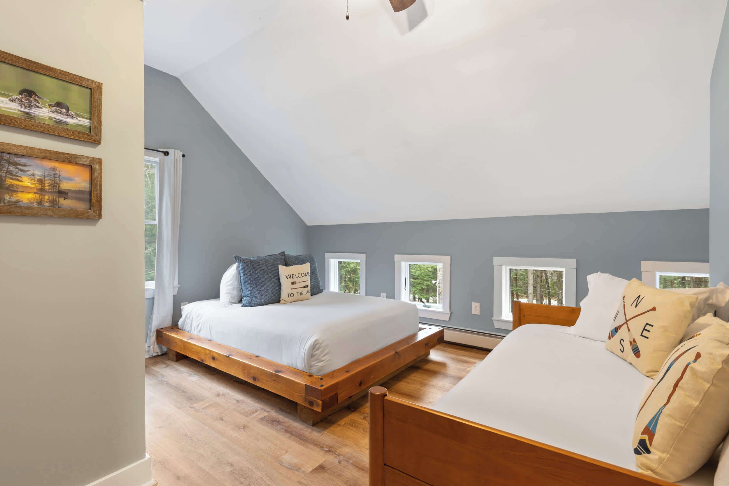 A bedroom with a sloped ceiling, a wooden bed frame, a white bed, and two windows with curtains.
