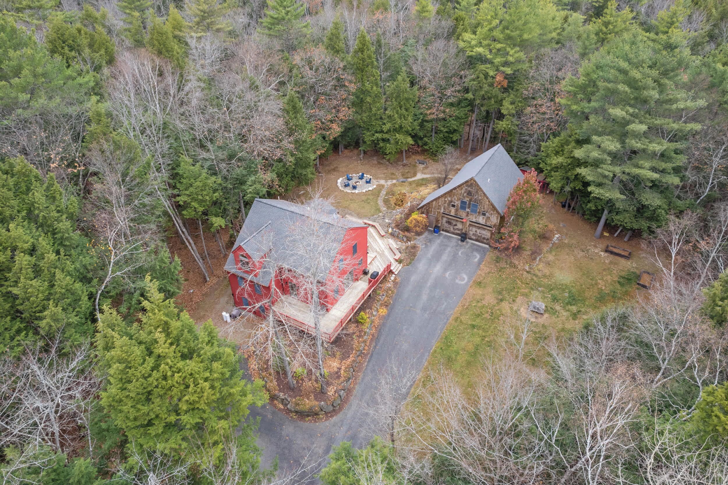 An aerial view of a cabin surrounded by dense forest, with a paved driveway leading to the entrance.