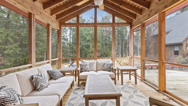 Un área de estar techada al aire libre con techo de madera, ventanas amplias y vistas al bosque.