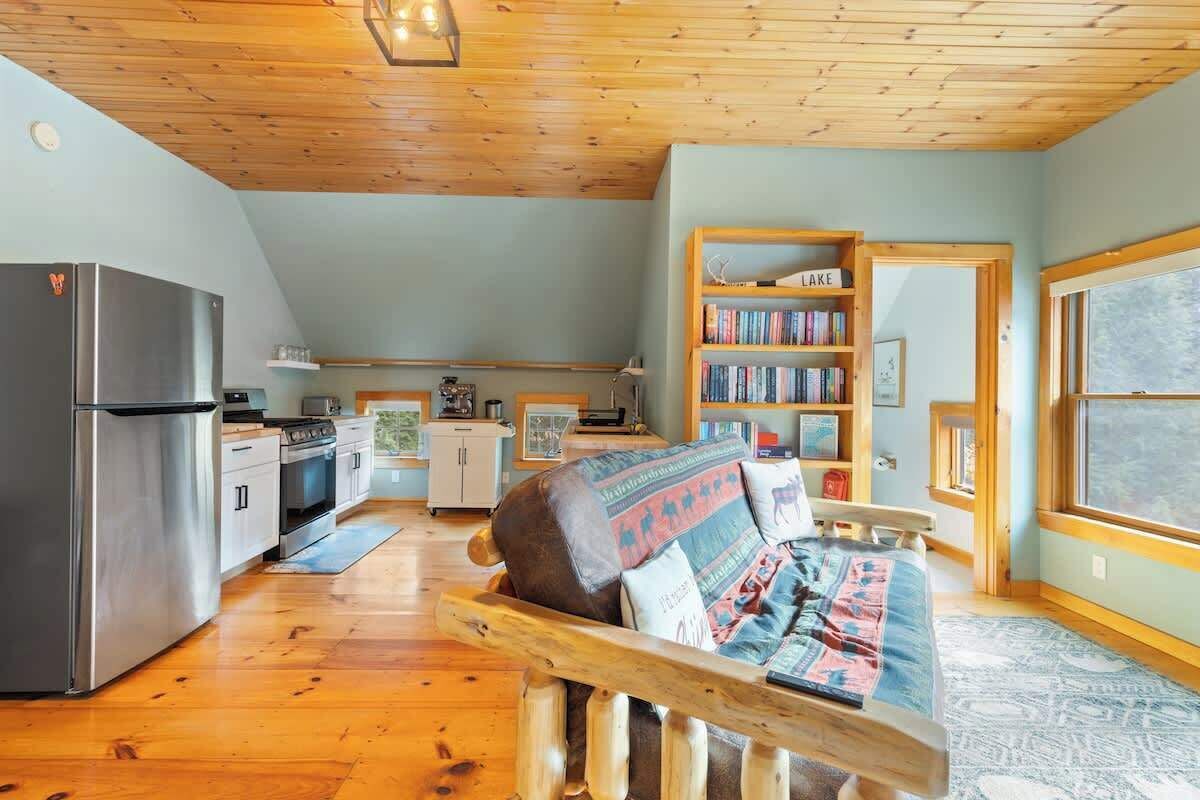 Un acogedor interior de cabaña con techo de madera, una kitchenette, un sofá con cojines estampados y una estantería.
