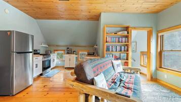 Un acogedor interior de cabaña con techo de madera, una kitchenette, un sofá con cojines estampados y una estantería.