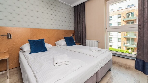 A hotel room with two beds, a window with a view of buildings, a radiator, and a wooden floor.