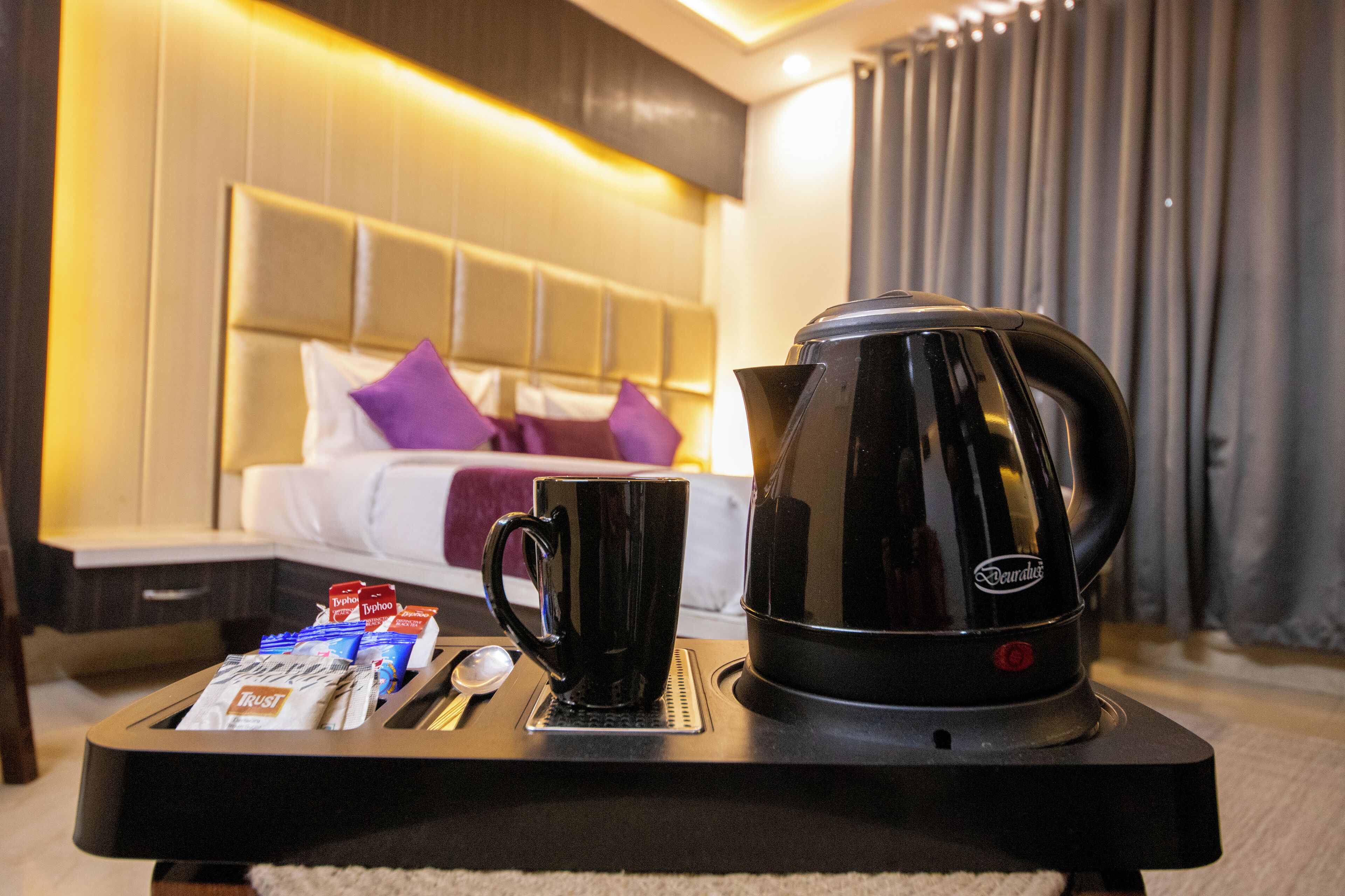A hotel room with a bed, a coffee maker, a mug, and some breakfast items on a tray.