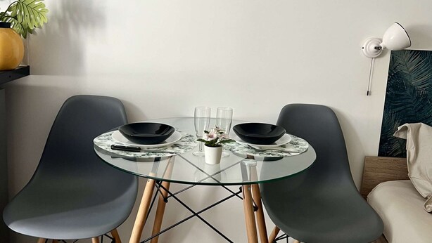 Un espace repas moderne avec une table en verre, deux chaises noires et un mur blanc orné d’un miroir et d’un tableau.