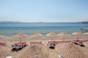 A beach with rows of white lounge chairs and woven umbrellas, a clear blue sea, and distant landforms.