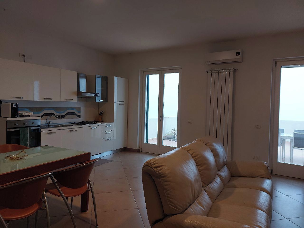 Una sala de estar con un sofá, un comedor con mesa y sillas, una cocina con armarios y electrodomésticos, y ventanas grandes con cortinas.