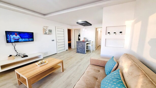Ein modernes Wohnzimmer mit einem Flachbildfernseher, einem Holztisch, einem beigefarbenen Sofa mit gemusterten Kissen und einer Küche im Hintergrund.