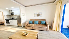 A compact living space with a kitchenette, a sofa with patterned cushions, a wooden coffee table, and a blue-tiled floor.