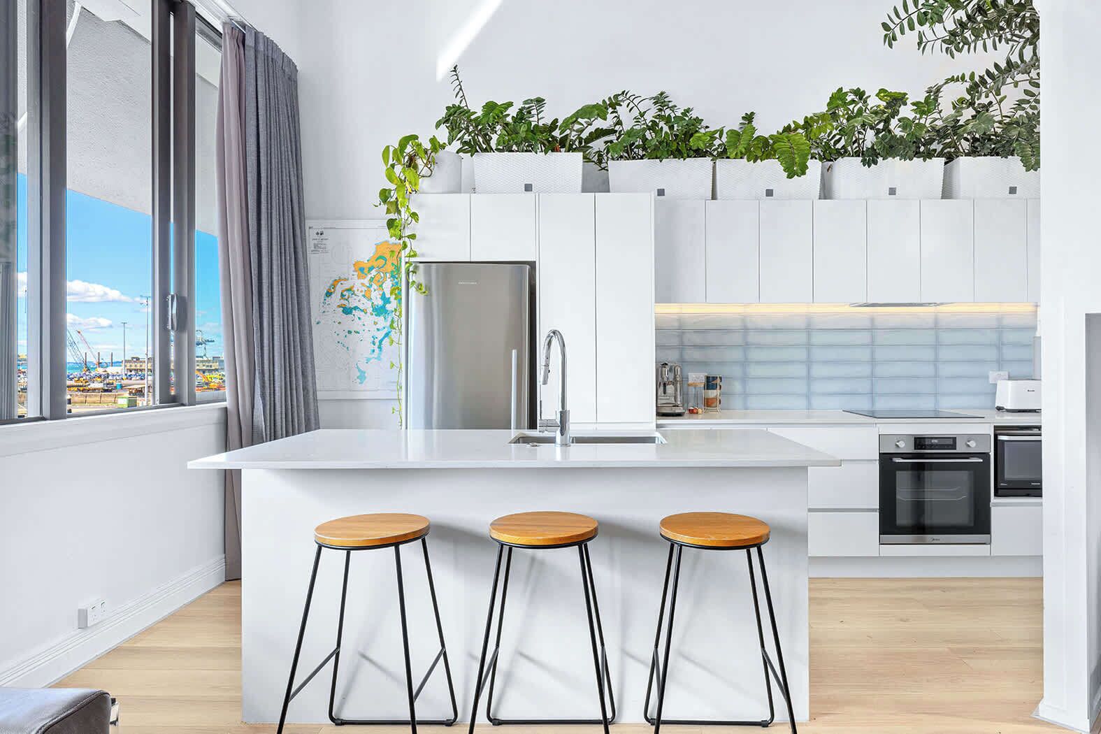 A modern kitchen with a central island, three stools, a refrigerator, an oven, and a window with a view of a construction site.