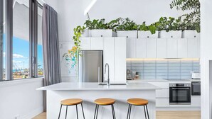 A modern kitchen with a central island, three stools, a refrigerator, an oven, and a window with a view of a construction site.