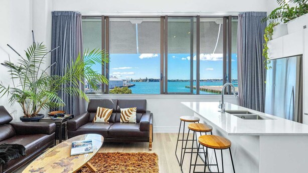 A modern living room with a sofa, coffee table, and bar stools. A large window offers a view of the sea and a pier.