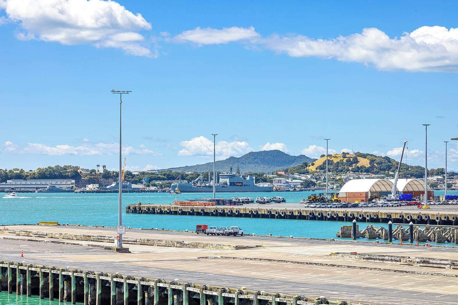 A coastal port with a large ship docked, a pier with vehicles, and a mountainous backdrop.