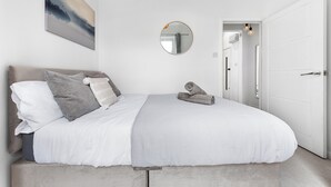 Een slaapkamer met een bed, kussens, een nachtkastje, een spiegel en een deur naar een andere kamer.