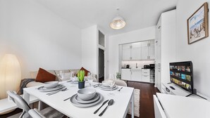 Een moderne eethoek met een witte tafel gedekt voor vier personen, een bank met kussens, een keuken op de achtergrond en een televisie aan de rechterkant.