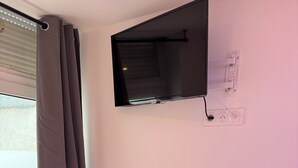 A small room with a TV mounted on the wall, a table, two chairs, a window with curtains, and a potted plant on the windowsill.
