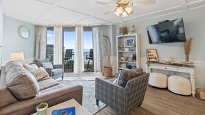 A living room with a sofa, armchair, and a TV on a white console. There is a ceiling fan, a window with curtains, and a view of the outdoors. - Follow the Moon Villa, St Regis Oceanfront in Topsail (North Topsail Beach)