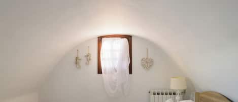 A bedroom with a sloped ceiling, a bed with a wooden headboard, a bedside table with a lamp, and a window with a sheer curtain.