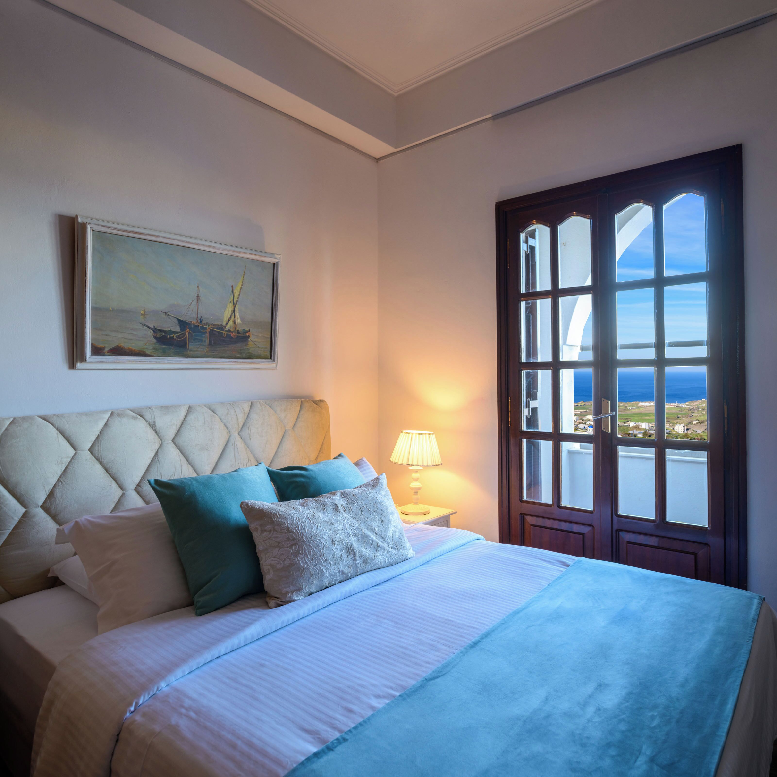 Una camera da letto con un letto, cuscini, un quadro, un comodino con una lampada e una finestra vista.
