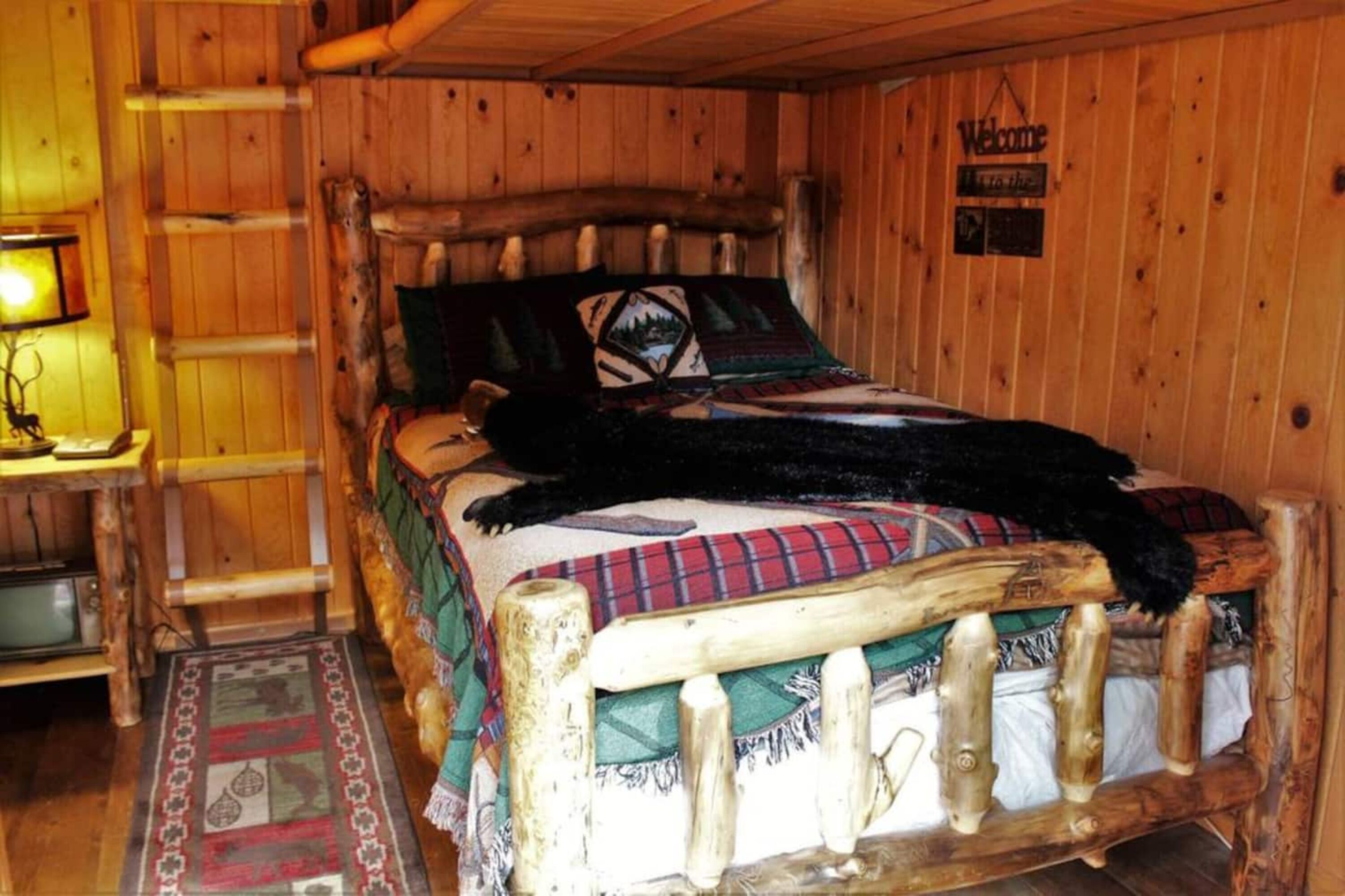 A wooden cabin interior with a bed featuring a blanket and pillows, a wooden ladder, and a welcome sign.